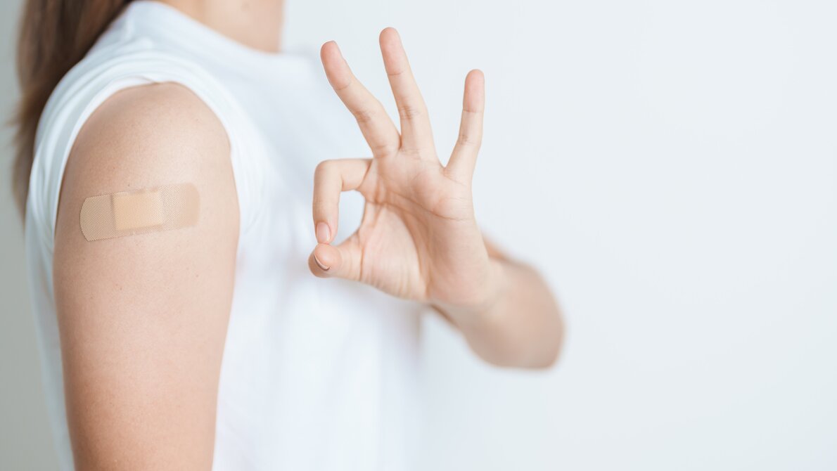 Das Bild zeigt einen Arm mit einem Pflaster, das auf eine kürzlich erfolgte Grippeimpfung  hinweist. Die Hand daneben formt ein "Okay"-Zeichen, was eine positive Botschaft vermittelt.