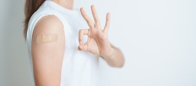 Das Bild zeigt einen Arm mit einem Pflaster, das auf eine kürzlich erfolgte Grippeimpfung  hinweist. Die Hand daneben formt ein "Okay"-Zeichen, was eine positive Botschaft vermittelt.