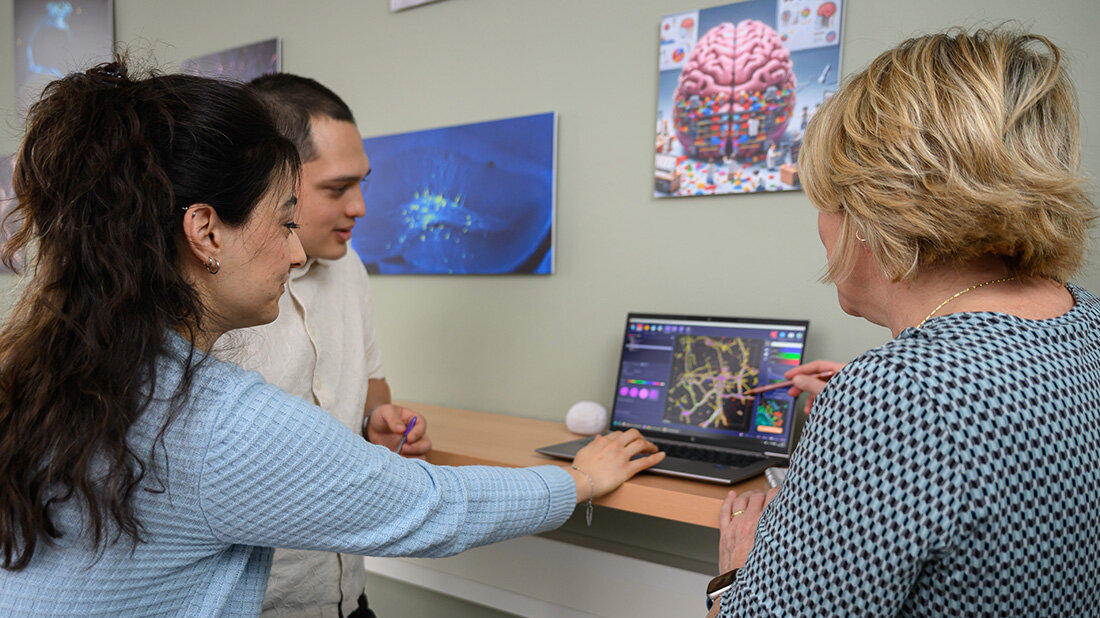 Drei Personen diskutieren vor einem Laptop mit wissenschaftlichen Visualisierungen, im Hintergrund hängen Bilder mit neurologischen und biologischen Motiven.