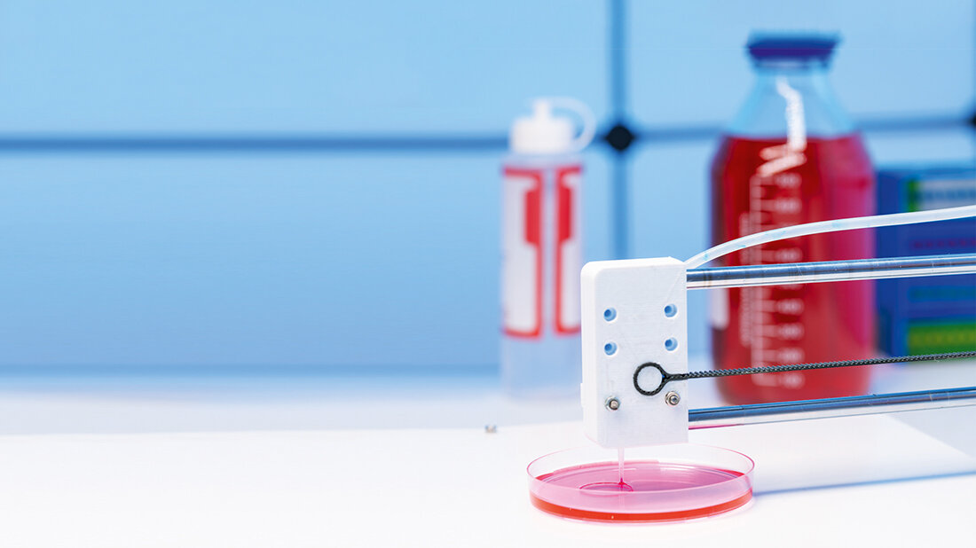 Ein 3D-Druckkopf gibt in einem Labor eine rosafarbene Flüssigkeit in eine Petrischale ab, während im Hintergrund unscharf Flaschen und Gefäße mit Nährlösungen zu sehen sind.