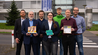 Eine Gruppe von 7 Personen einer Forschungsgruppe vor einem Gebäude mit Modellen in den Händen, Fahne der Hochschule Koblenz im Hintergrund
