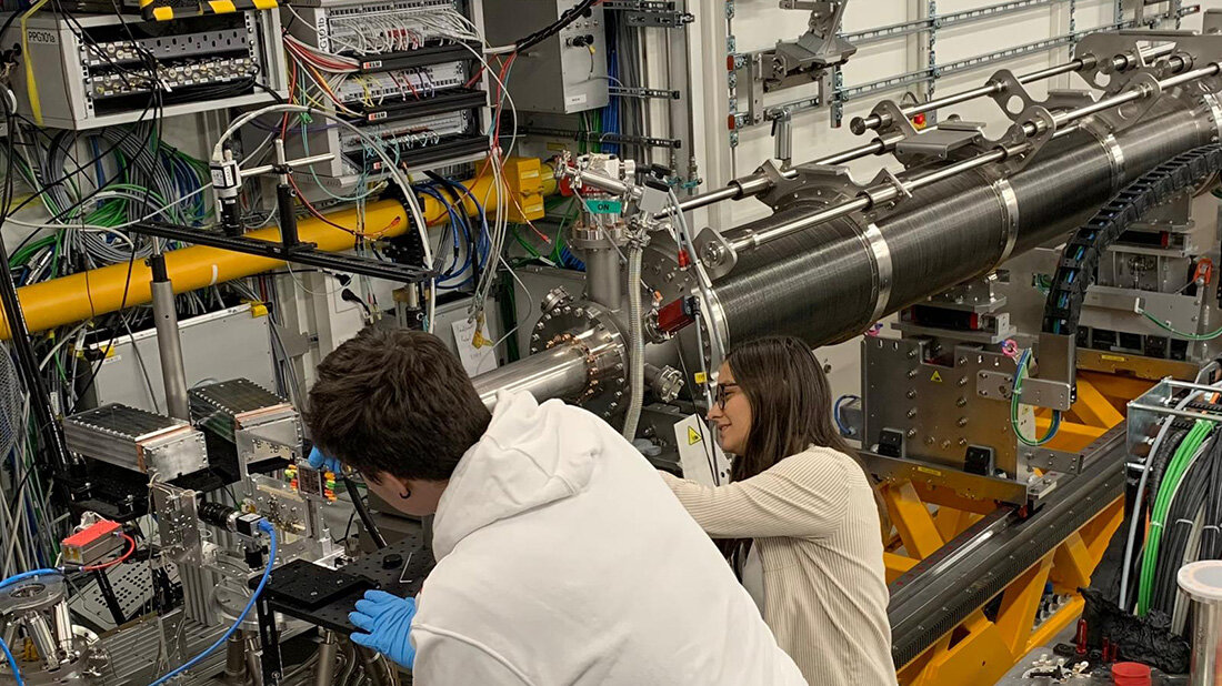 Zwei Personen arbeiten an einer komplexen wissenschaftlichen Apparatur in einem Labor, umgeben von Kabeln, einem größeren Rohr, Maschinen und technischen Geräten.