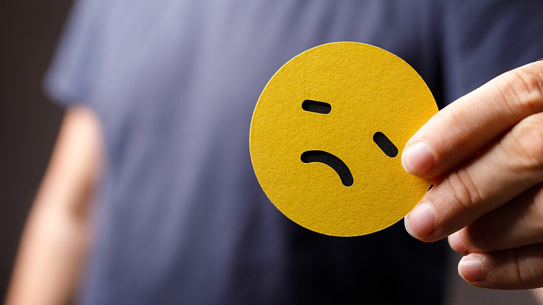 Ein trauriger, gelber Smiley wird von einer Hand gehalten, Oberkörper einer Person unscharf im Hintergrund