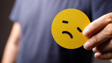 Ein trauriger, gelber Smiley wird von einer Hand gehalten, Oberkörper einer Person unscharf im Hintergrund