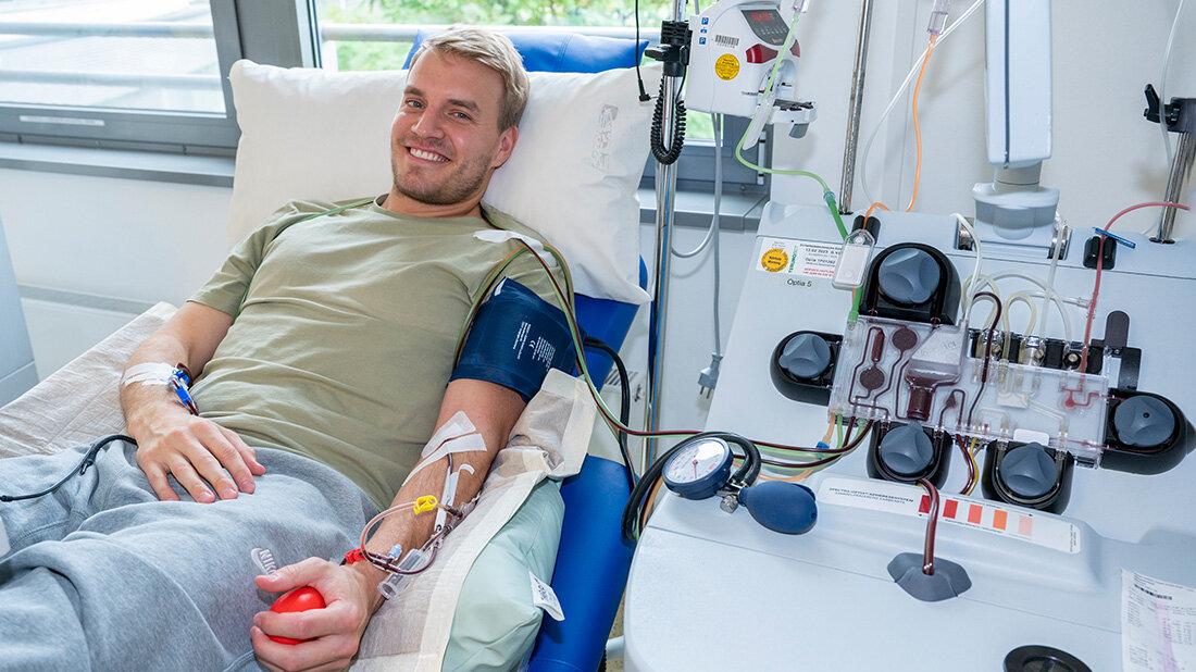 Stammzellspende.jpg Mann liegt auf einer Praxisliege, spendet Blut und drückt mit der linken Hand einen Ball.