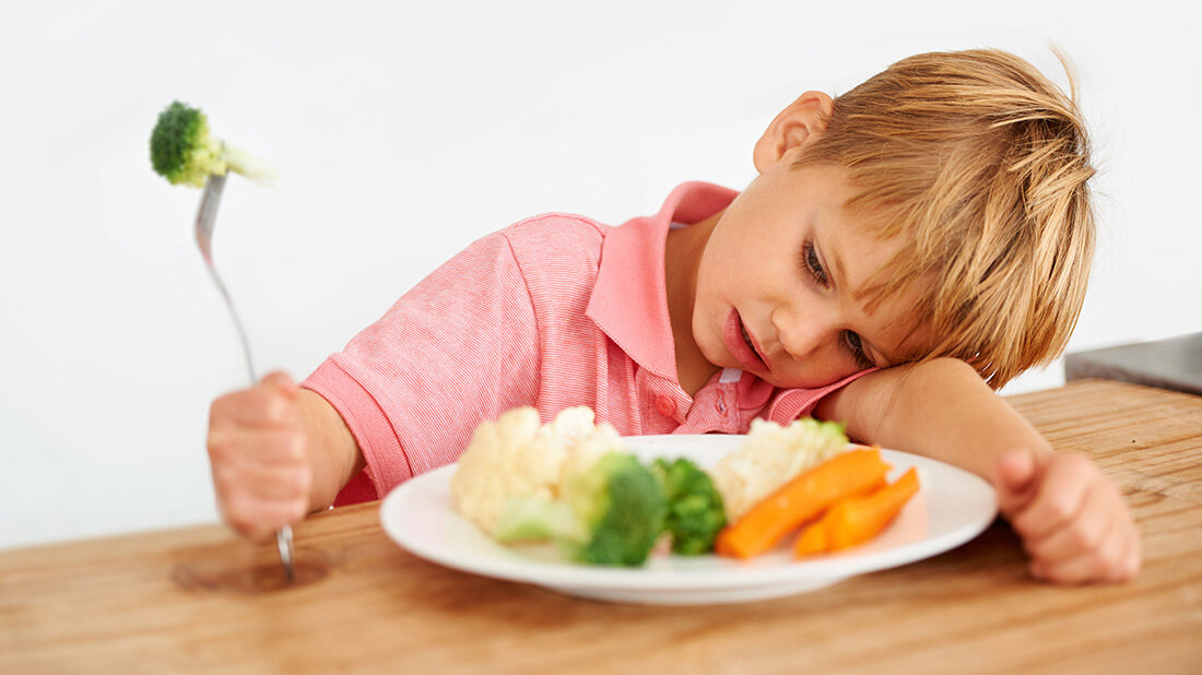 Junge sitzt am Tisch mit Gemüse auf einem Teller vor sich, Gabel in der Hand, Kopf auf dem Arm abgelegt