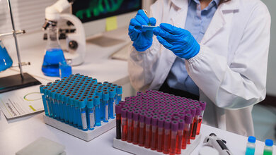  Wissenschaftlerin im Labor untersucht eine Probe, umgeben von Reagenzgläsern mit blauer und roter Flüssigkeit, Mikroskop im Hintergrund.