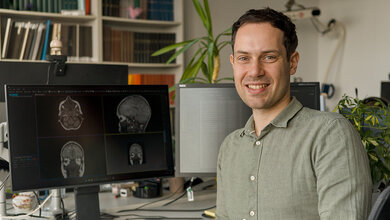 Mann in Büro am Schreibtisch, auf einem Bildschirm hinter ihm sind MRT-Aufnahmen zu sehen, Blumen und Bücherregale im Hintergrund.