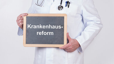 Ein Mediziner in weißem Kittel hält eine Schiedertafel vor die Brust, auf die Krankenhausreform geschrieben ist