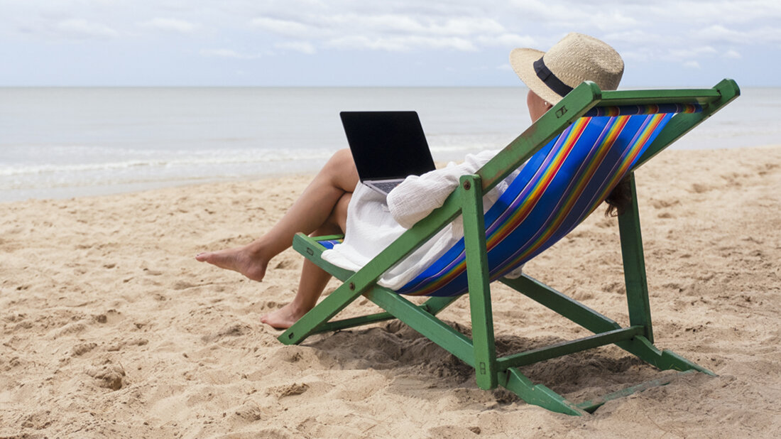  Das Bild zeigt eine Person, die in einem Liegestuhl am Strand sitzt und einen Laptop benutzt.