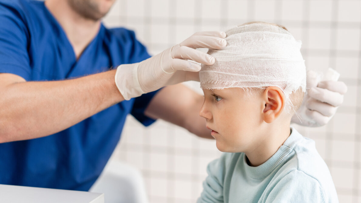 Das Bild zeigt eine medizinische Situation, in der ein Kind von einem Fachmann behandelt wird. Der Arzt trägt Handschuhe und legt einen Verband um den Kopf des Kindes, was auf eine Verletzung oder eine postoperative Versorgung hinweist.