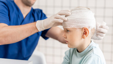 Das Bild zeigt eine medizinische Situation, in der ein Kind von einem Fachmann behandelt wird. Der Arzt trägt Handschuhe und legt einen Verband um den Kopf des Kindes, was auf eine Verletzung oder eine postoperative Versorgung hinweist.