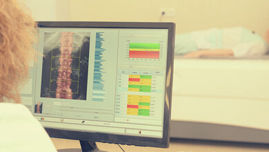 Bildschirm zeigt eine medizinische Analyse der Wirbelsäule, im Hintergrund liegt eine Patientin auf einer Untersuchungsliege.