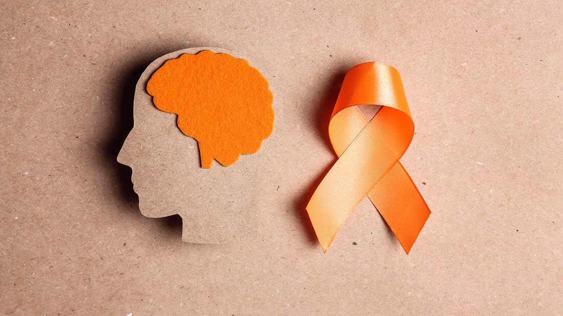 Multiple_Sklerose_orange_Schleife.jpg Silhouette eines Kopfes mit orangefarbenem Gehirn aus Filz neben einer orangefarbenen Schleife auf braunem Hintergrund, symbolisiert Bewusstsein für MS.