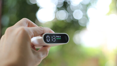 Eine Hand hält einen Partikelsensor PM2,5 mit dem Wert 013 vor einem verschwommenen Naturhintergrund