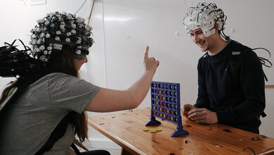 Probanden einer medizinischen Studie beim Vier-gewinnt-Spiel ab einem Holztisch mit Messsonden am Kopf.