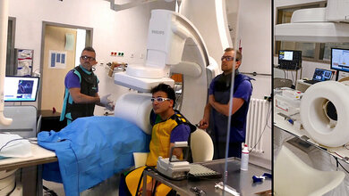 Medizinisches Personal in einem Untersuchungsraum mit moderner Bildgebungs- und Diagnosetechnik, Patient in Schutzkleidung unter einem Gerät sitzend für eine MPI-Untersuchung.