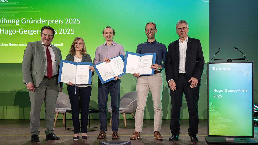 Preisverleihung_GruenderpreisBayern.jpg Fünf Personen auf einer Bühne bei einer Preisverleihung, Bildschirm mit blau/grünem Farbverlauf