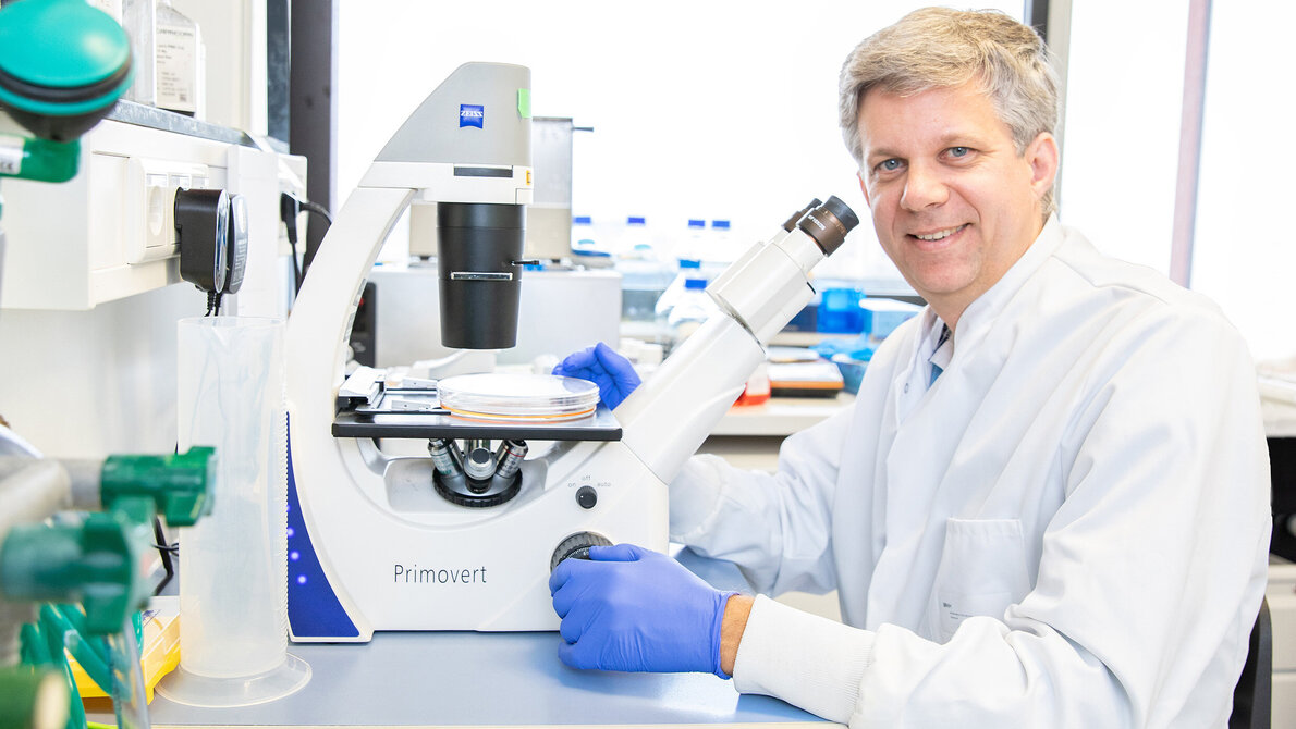 Ein Mann (Professor Dölken) in weißem Kittel sitzt an einem Tisch vor einem Mikroskop im Labor.