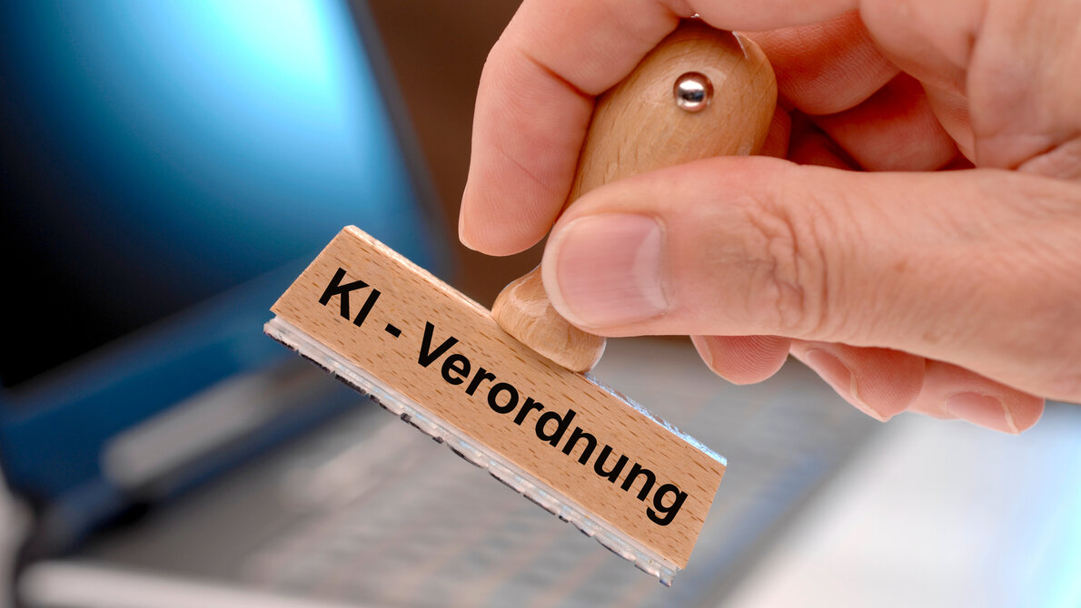 Das Bild zeigt einen Holzstempel mit der Aufschrift „KI - Verordnung", der von einer Hand gehalten wird. Im Hintergrund ist ein Laptop zu sehen, was auf den Zusammenhang zwischen Künstlicher Intelligenz (KI) und rechtlichen Regelungen hinweist.