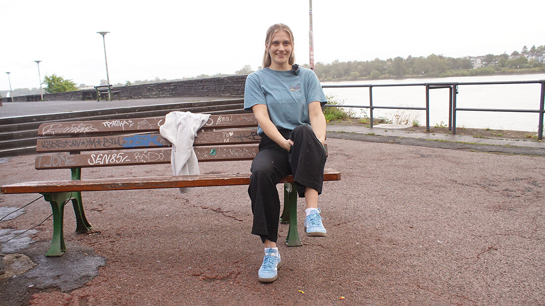 Frau sitzt auf einer mit Graffiti bedeckten Parkbank am Flussufer, trägt ein blaues T-Shirt, schwarze Hose und blaue Turnschuhe, daneben liegt ein grauer Pullover.