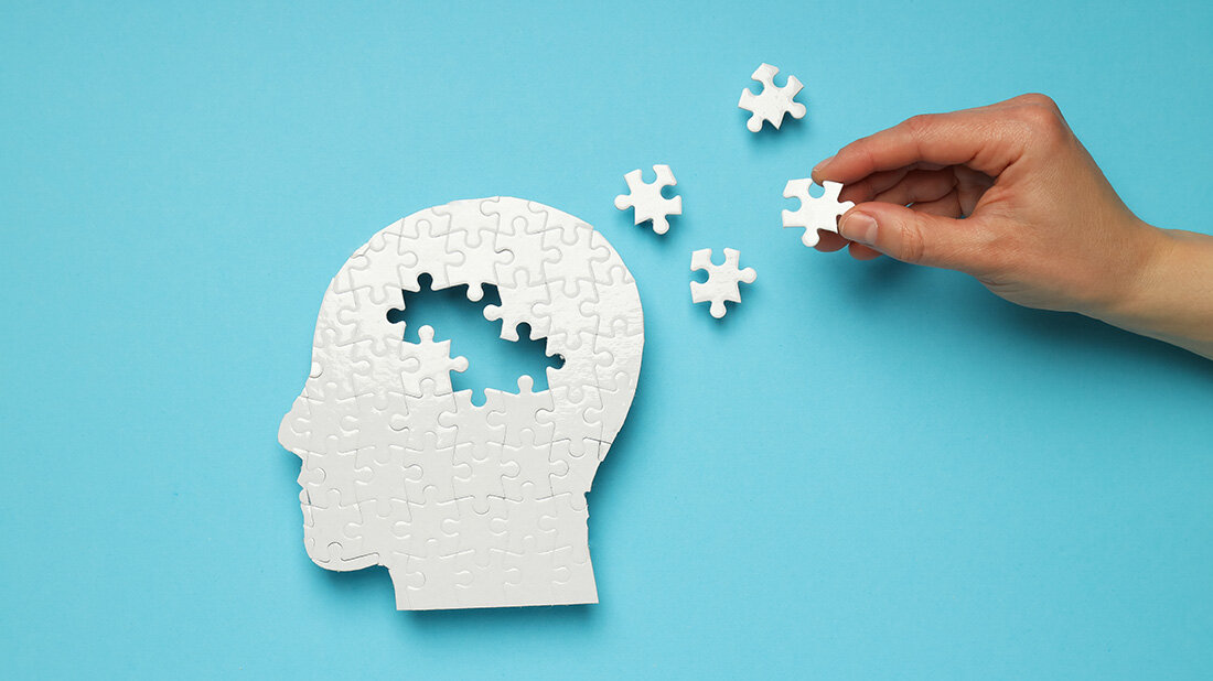 Weißes Puzzle eines menschlichen Kopfes auf blauem Hintergrund mit vier fehlenden Puzzleteilen, die daneben liegen und eine Hand hält ein Teil fest.