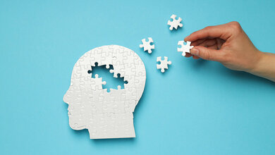 Weißes Puzzle eines menschlichen Kopfes auf blauem Hintergrund mit vier fehlenden Puzzleteilen, die daneben liegen und eine Hand hält ein Teil fest.