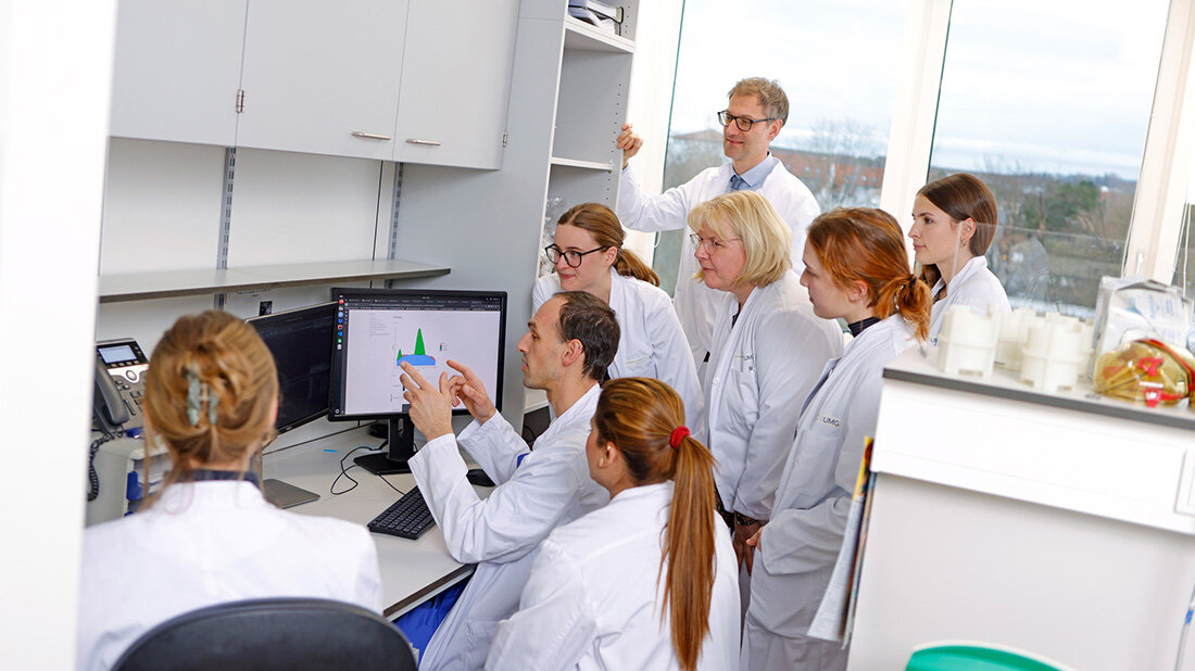 Mehrere Personen in weißen Kitteln stehen vor einem Schreibtisch, während eine sitzende Person etwas erklärt.