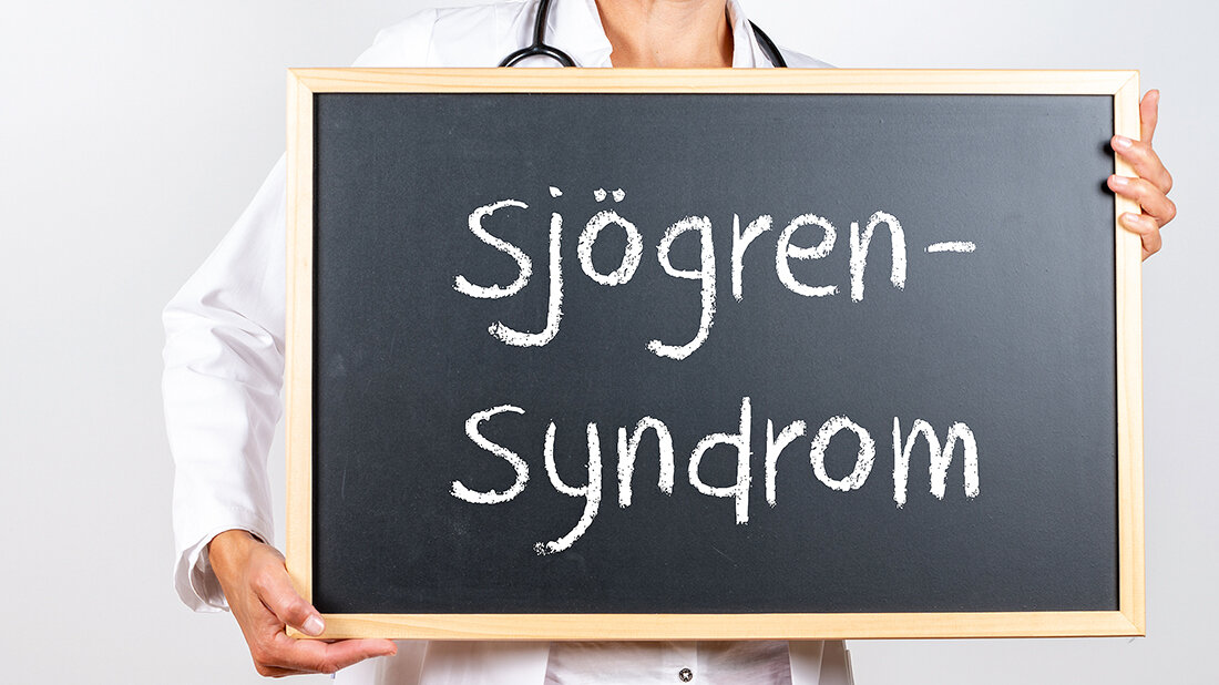 Eine Person im weißen Kittel mit Stethoskop hält sich eine Tafel mit dem Schriftzug „Sjögren-Syndrom“ vor die Brust