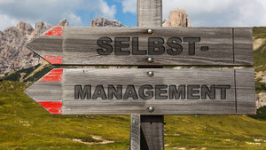 Wegweiser vor einem Hochgebirgshintergrund, auf denen das Wort „SELBSTMANAGEMENT“ steht