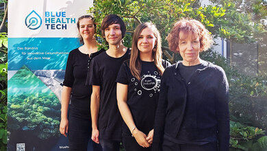 Pardemann_Pfeifer_Mueller_Classen.jpg Vier Personen in schwarzer Kleidung vor einem Banner mit der Aufschrift "Blue Health Tech" und grüner Pflanzenkulisse im Hintergrund.