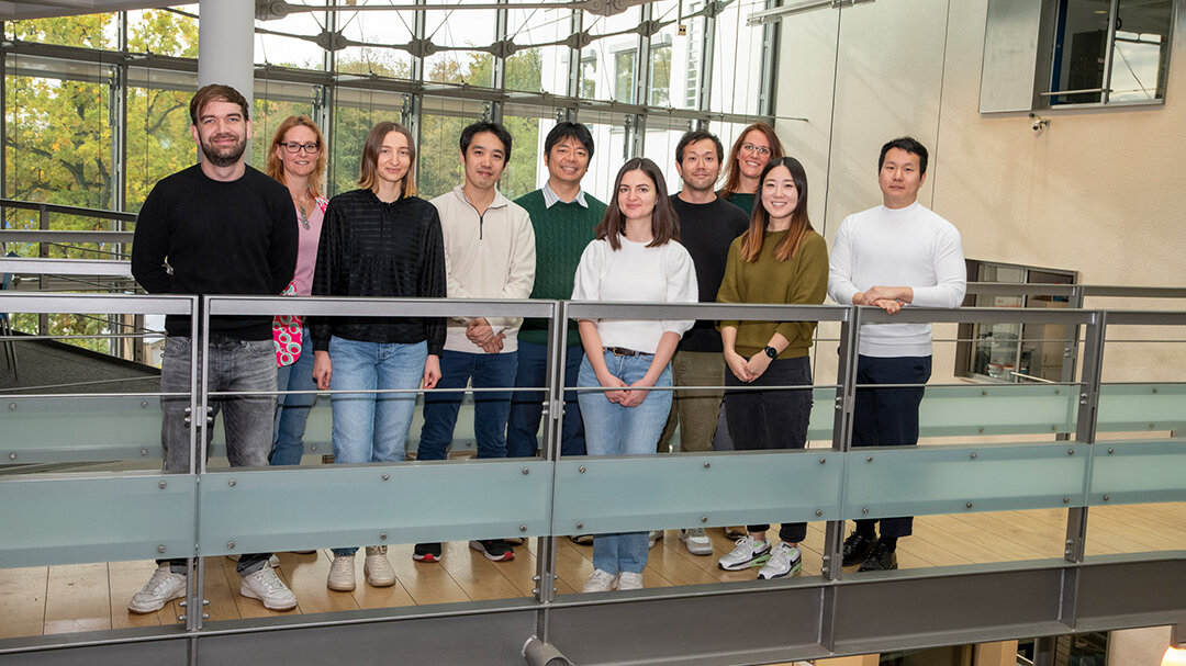 Gruppenfoto mit verschiedenen Menschen auf einer gläsernen Galerie, im Hintergrund ist eine große Glasfront, durch die Bäume zu sehen sind.