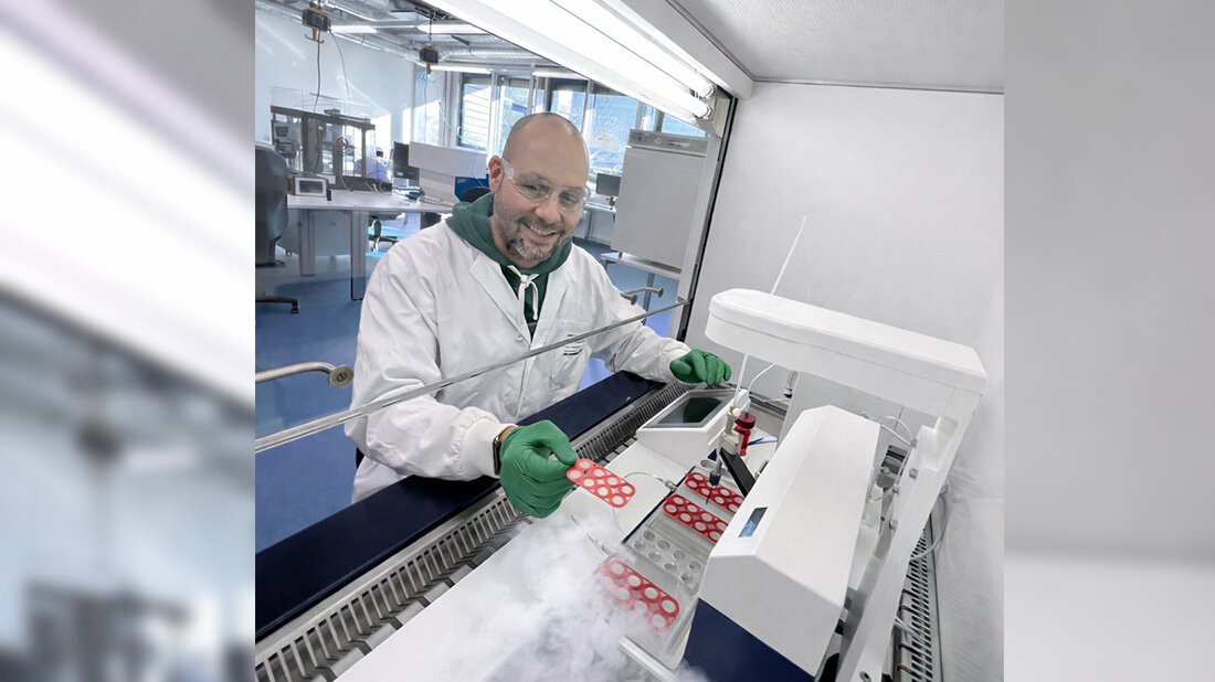 Kryokonservierung_Fraunhofer.jpg Laborant in Schutzkleidung arbeitet an einer Maschine mit roten Proben in einer sterilen Umgebung. Im Vordergrund sind Probenbehälter und etwas wie Trockeneis sichtbar.