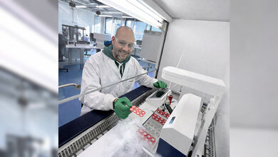 Laborant in Schutzkleidung arbeitet an einer Maschine mit roten Proben in einer sterilen Umgebung. Im Vordergrund sind Probenbehälter und etwas wie Trockeneis sichtbar.