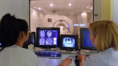 Zwei medizinische Fachkräfte überwachen MRT-Scans auf Monitoren in einem Kontrollraum, während ein Patient im MRT-Gerät liegt.