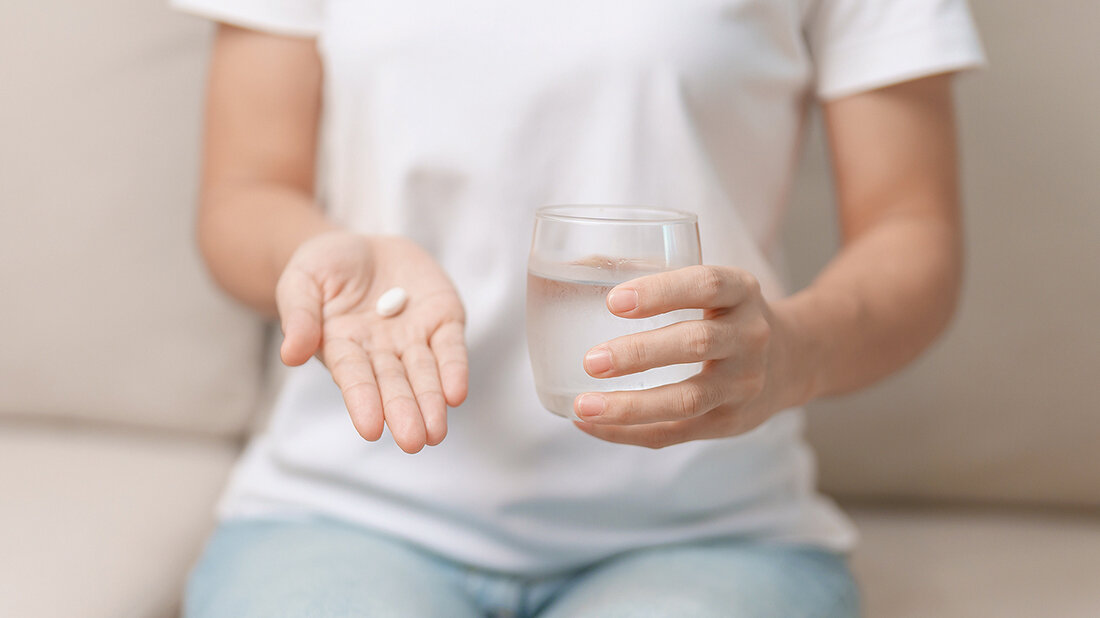 Foto, auf dem eine Person in der flachen rechten Hand eine Tablette liegen hat und mit linken ein Glas Wasser hält