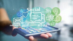 Eine Hand hält ein Smartphone, über dem Symbole und Begriffe zu Social Media und digitaler Kommunikation schweben