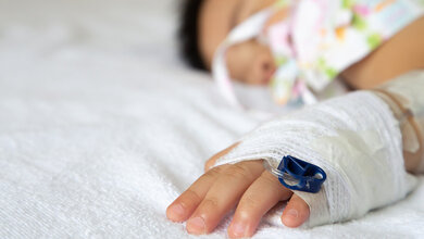 Hand eines Babys mit einem Verband und einem Zugang liegt auf einem Bett, Gesicht des Babys ist verschwommen im Hintergrund zu sehen