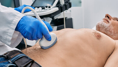 Bauchultraschall.jpg Ein Arzt führt eine Bauchultraschalluntersuchung am Oberkörper eines männlichen Patienten durch, das Ultraschallgerät ist im Hintergrund.