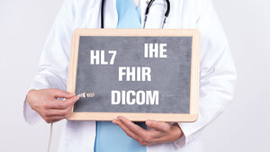 Eine Person im Arztkittel mit umgehängtem Stethoskop hält eine Schiefertafel vor der Brust, auf der die Kürzel „HL7“, „IHE“, „FHIR“ und „DICOM“ zu lesen sind
