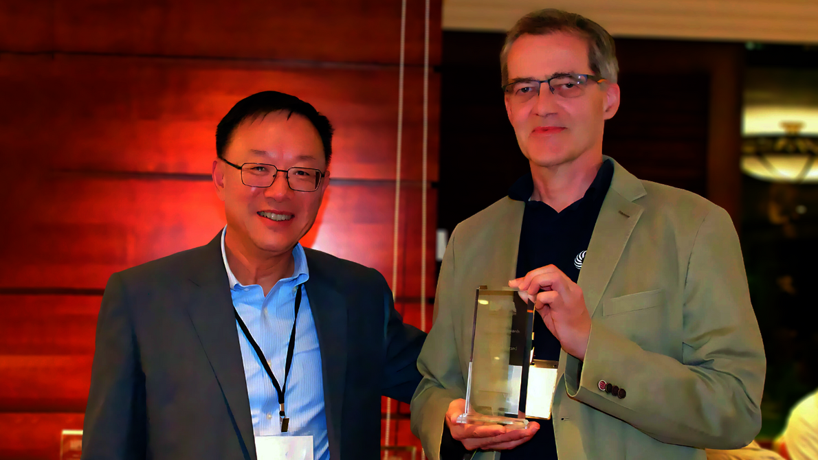 Zwei Männer, der eine legt den Arm um den anderen, der einen Preis in der Hand hält und präsentiert.