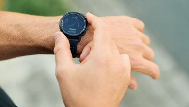 Person überprüft Daten auf einer Smartwatch, die rechte Hand drückt seitlich auf die Uhr an der linken Hand, grauer unscharfer Hintergrund.