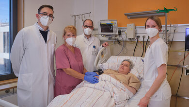 Medizinisches Team in einem Krankenhauszimmer, das eine Patientin am Krankenbett betreut, umgeben von medizinischen Geräten und Monitoren.