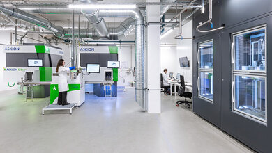Heidelberg-cell-and-liquid-biobank.jpg Moderne Laborumgebung mit technischen Geräten und Arbeitsplätzen. Eine Frau in Laborkleidung arbeitet an einem Gerät, während ein Mann im Hintergrund am Computer sitzt.