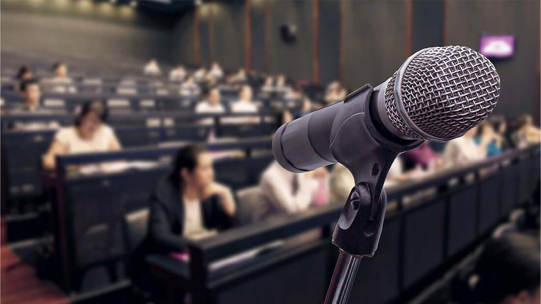 Großaufnahme eines Mikrofons, im Hintergrund ein Hörsaal