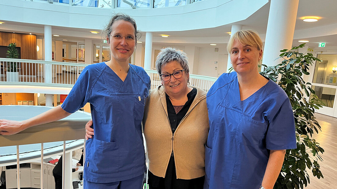 Drei Personen stehen in einem modernen, hellen Gebäude. Zwei tragen blaue medizinische Kleidung, die mittlere Person trägt eine beige Strickjacke.
