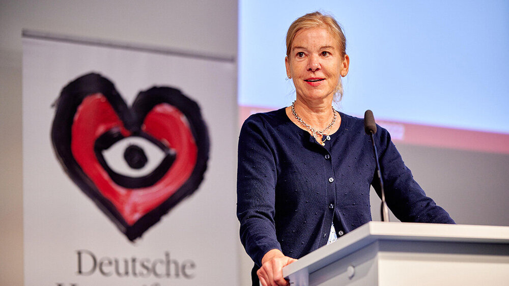 Frau an einem Podium, im Hintergrund Grafik eines Herzens mit einem Auge drin