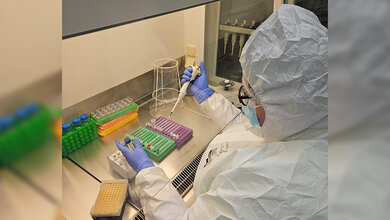 Labortechnikerin in weißem Schutzanzug mit Pipette in der Hand und Proben vor sich.
