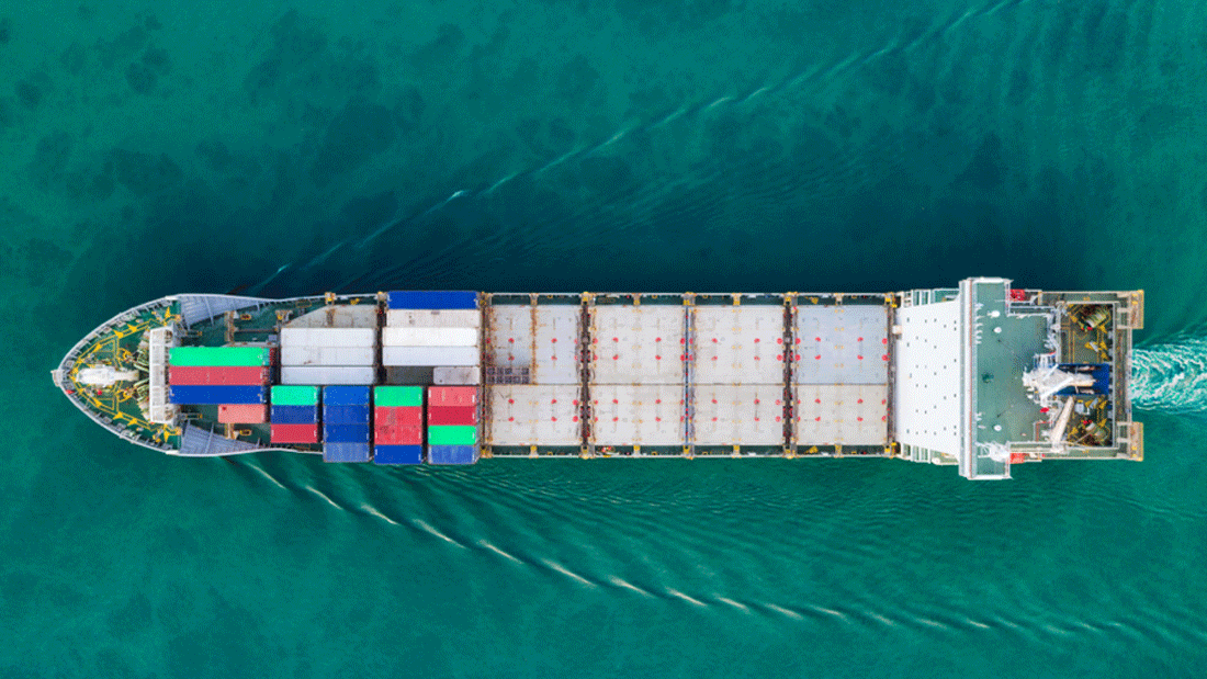 Dieses Bild zeigt eine Luftaufnahme eines Frachtschiffs, das durch klares, blaues Wasser fährt. Das Schiff transportiert mehrere bunte Container, die vermutlich für den Gütertransport verwendet werden. 
