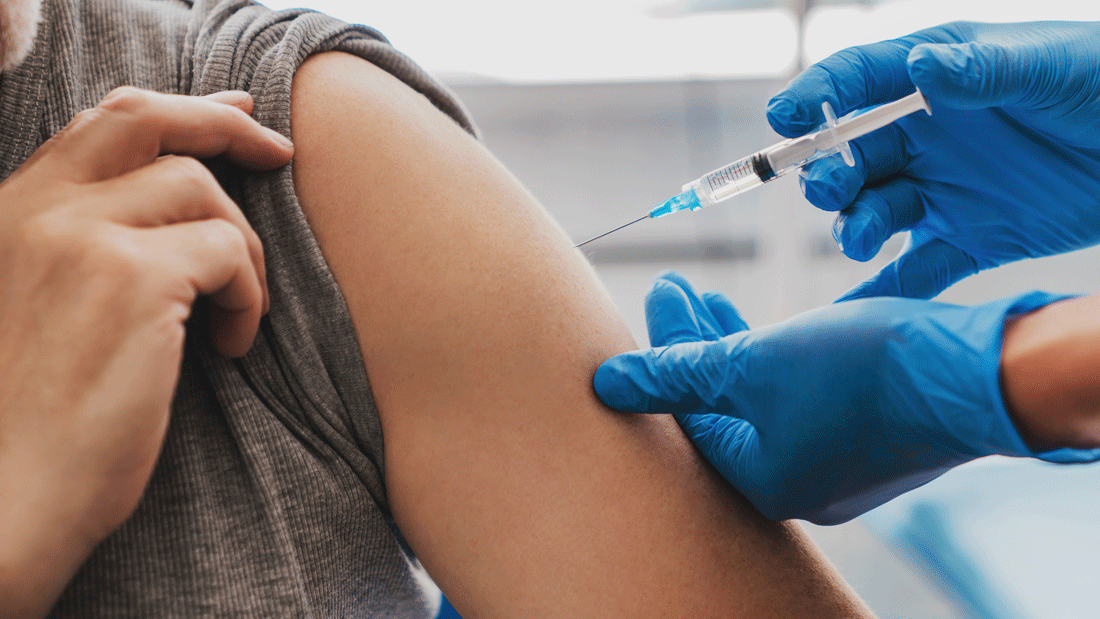 Nahaufnahme einer Injektion in den Oberarm: Eine Person zieht den Ärmel hoch, während medizinisches Personal mit blauen Handschuhen eine Spritze ansetzt.
